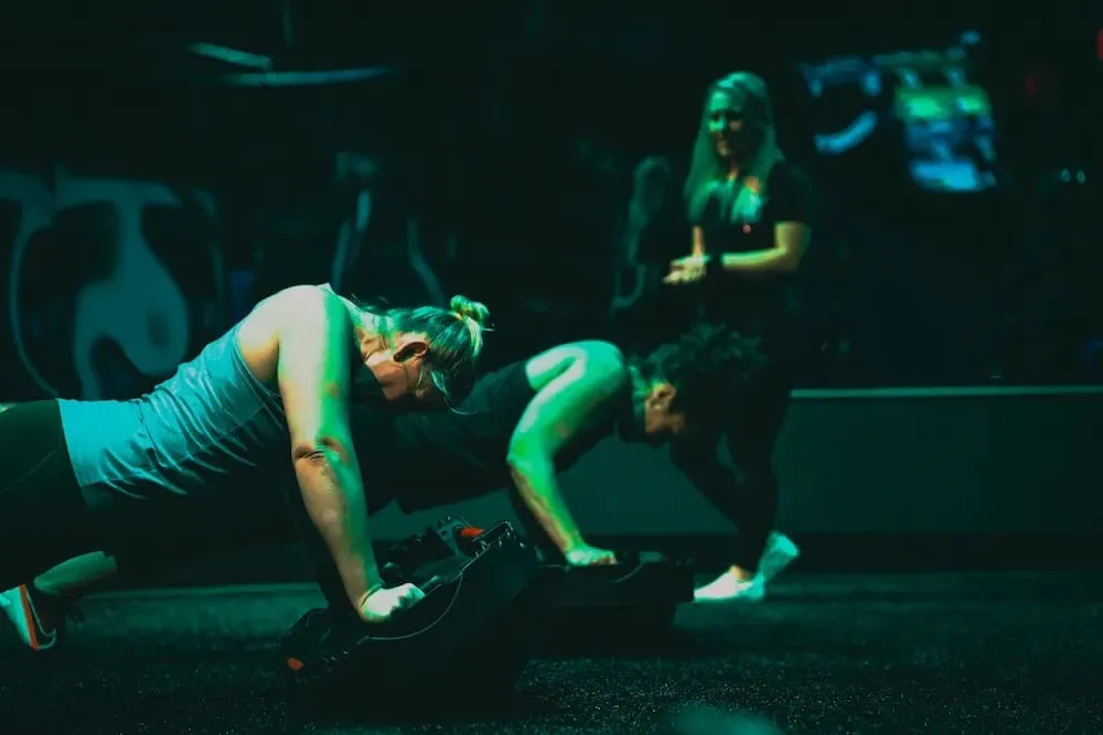 A woman performs a landmine squat in a dimly lit gym, holding a barbell anchored to the floor while a man exercises in the background near weight racks.